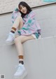 A woman sitting on a wall wearing a tie dye sweatshirt and white shorts.