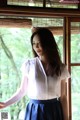 A woman standing in front of a window wearing a white shirt and blue skirt.