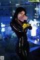 A woman in a black latex outfit holding a yellow tape around her mouth.