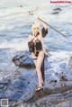 A woman in a bikini holding a spear on a rocky beach.